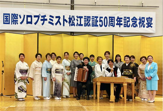 国際ソロプチミスト松江認証50周年記念祝宴にて、ツィンバロンの斉藤浩とアコーディオンの歌島昌智が松江の会員の皆さまと金屏風の前で記念撮影