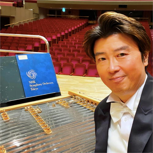 タキシード姿の斉藤浩さんがイール・ツィンバロンとNHK交響楽団の譜面台と共に微笑む。空席のコンサートホール。