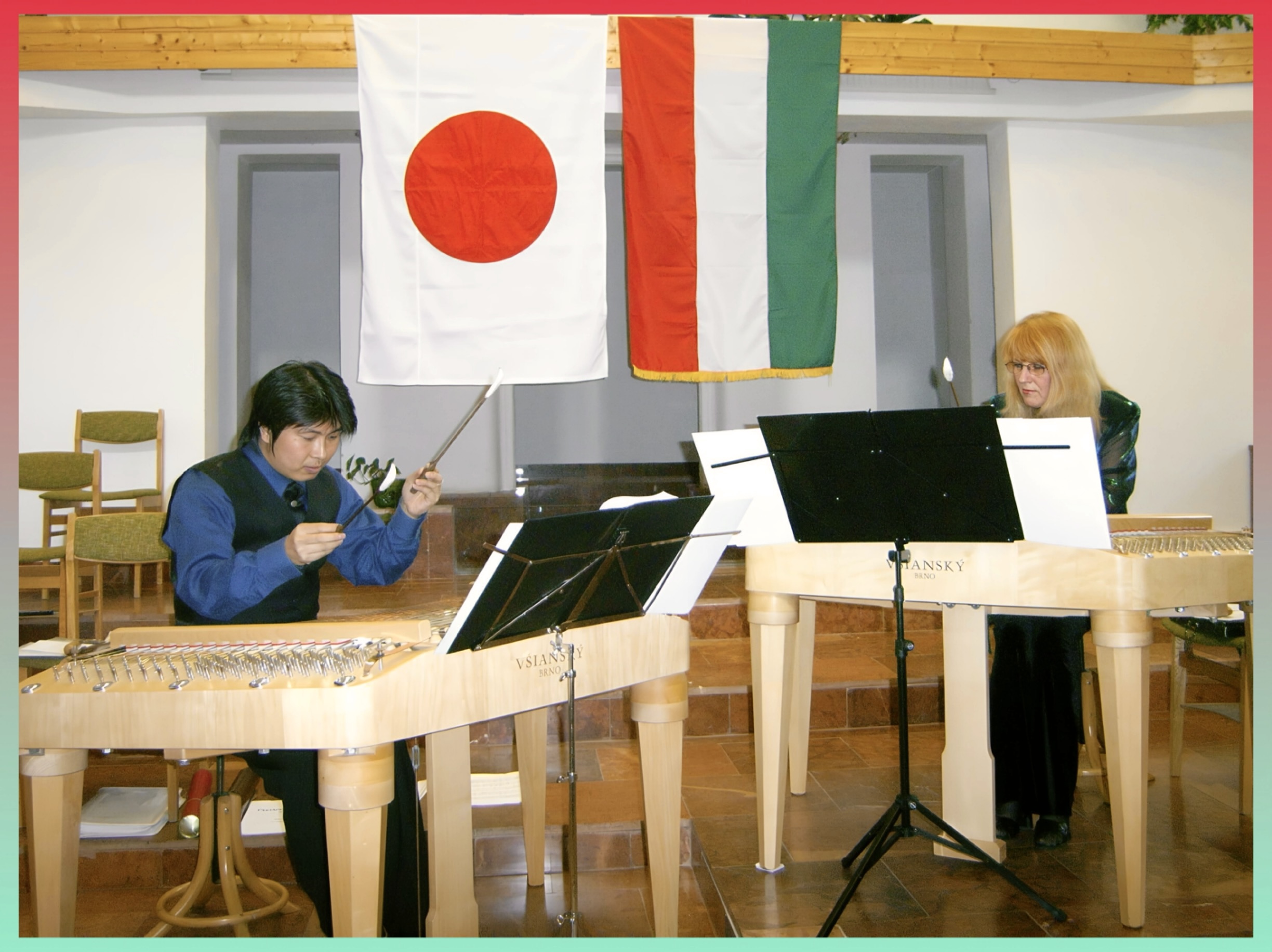 ブダペストのコンサートホール。背景の壁に日本の日の丸とハンガリー国旗が並んで掲げられている。舞台中央で2台の明るい木製のツィンバロンVSIANSKYが左右に置かれ、若い斉藤浩さんと金髪の女性が譜面台を挟んでツィンバロンを演奏している。斉藤さんは青いシャツに黒いベスト、手にバチを持ち真剣な表情。右側の女性はヘレンチャール・ヴィクトーリア師匠で、眼鏡をかけ濃い緑の衣装を着て楽譜を見つめている。床は大理石調、木の梁が見える温かい空間。