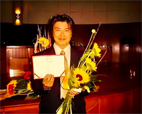 2006年ツィンバロンディプロマを取得した20年前の斉藤浩さん。黒スーツで証書とひまわりの花束を持ち、20年の歩みが始まった瞬間を笑顔で記録した一枚。